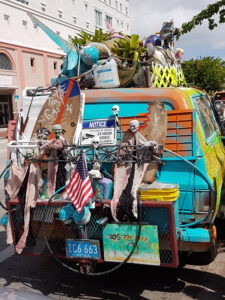 Ein bunt bemalter, mit Kunstobjekten beladener Lieferwagen in den Straßen von Miami, geschmückt mit skurrilen Dekorationen wie Skeletten, Flaggen und Pflanzen. Ein "Tips for Gas" Schild und eine amerikanische Flagge hängen an der Rückseite des Fahrzeugs.