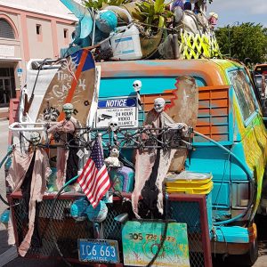 Ein bunt bemalter, mit Kunstobjekten beladener Lieferwagen in den Straßen von Miami, geschmückt mit skurrilen Dekorationen wie Skeletten, Flaggen und Pflanzen. Ein "Tips for Gas" Schild und eine amerikanische Flagge hängen an der Rückseite des Fahrzeugs.